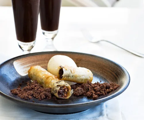 Plate of rolled pastries with chocolate filling, a scoop of ice cream, chocolate crumbs, and two dark drinks in the background.