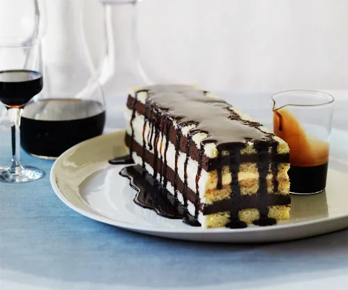Rectangular chocolate layer cake drizzled with chocolate sauce on a plate, with glasses of dark wine beside it.
