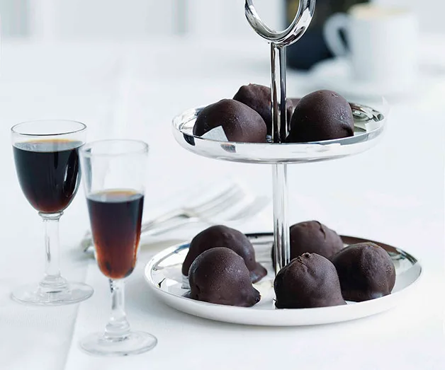 A two-tiered tray with chocolate truffles beside two glasses of dark dessert wine on a white tablecloth.