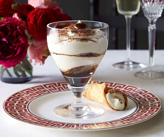 Glass of tiramisu layered dessert with cocoa dusting, on a decorative plate beside a cannoli and a vase of red flowers.