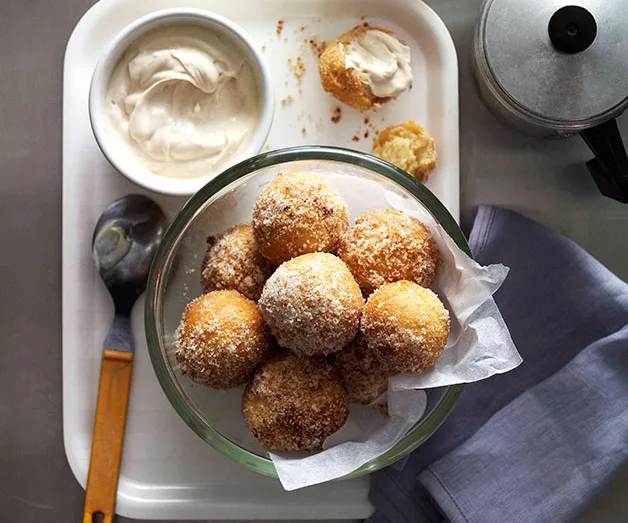 Sugared doughnuts in a bowl on a tray with dip, next to a coffee maker and cloth napkin.