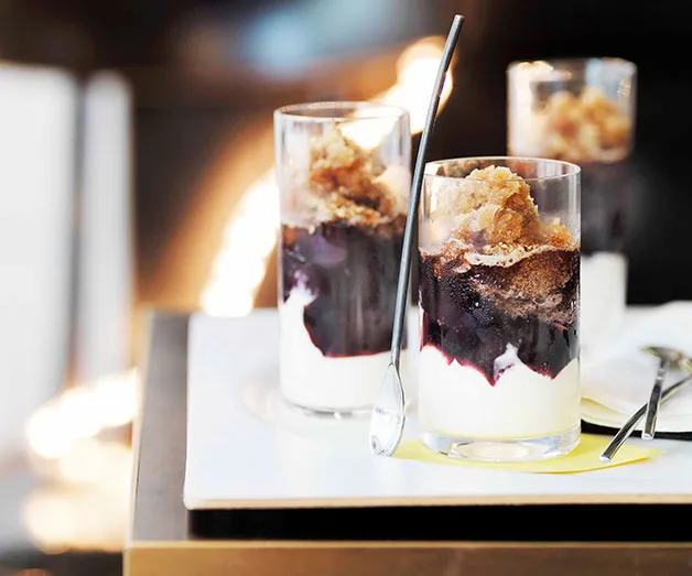 Three glass parfaits with layers of yogurt, berry compote, and crumble topping on a white tray, spoons placed beside.