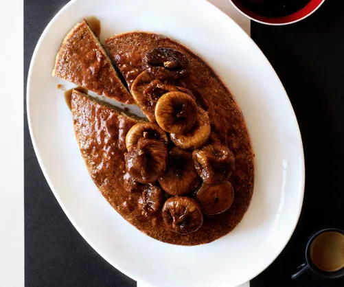 Oval-shaped cake with sliced figs on top, served on a white plate with a small black cup and red saucer nearby.