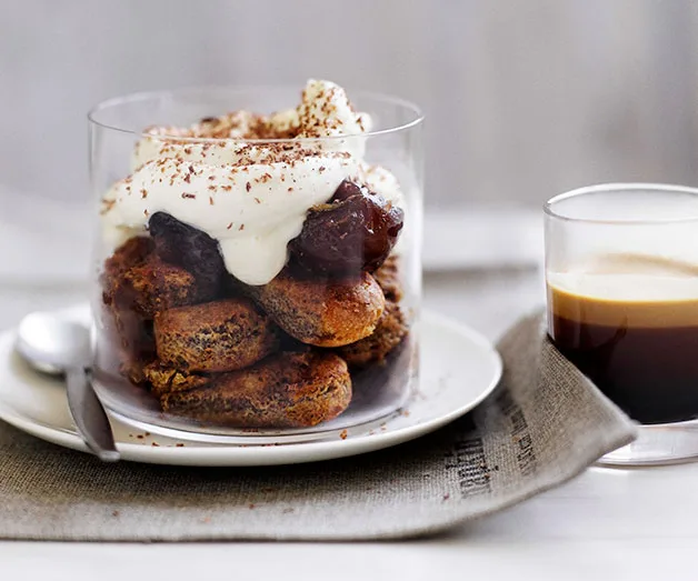 Tiramisu dessert in a glass, topped with cream and chocolate shavings, served beside a small cup of espresso on a napkin.
