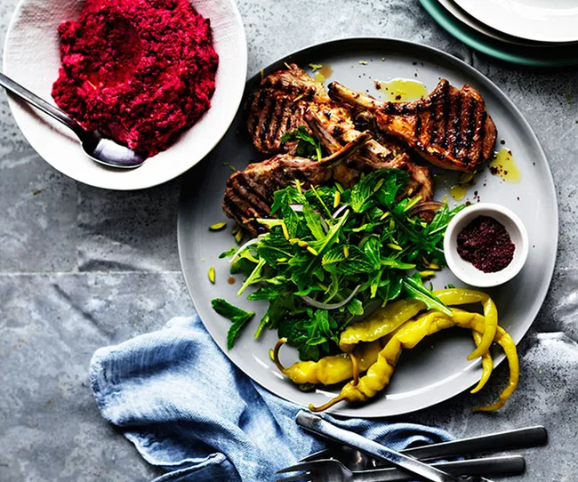 Grilled lamb chops with salad and peppers on a plate, served with beetroot dip and sumac, set on a textured surface.