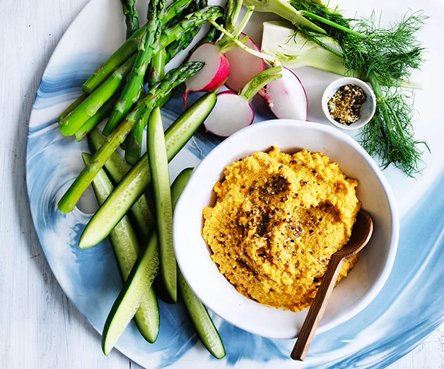 Plate of fresh vegetables with a bowl of yellow hummus, garnished with black pepper, on a blue and white plate.