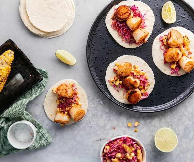 Scallop tacos with purple slaw and corn on tortillas, lime wedges, and grilled corn on a gray background.