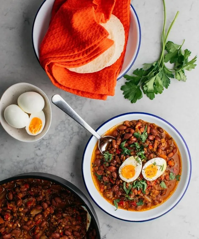 Bowl of bean stew topped with boiled eggs and herbs, served with tortillas on an orange cloth.