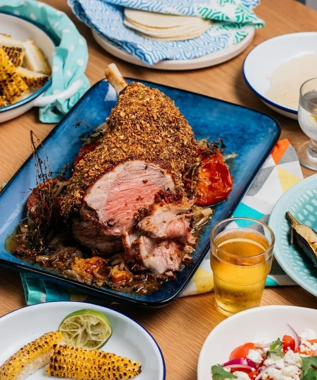 Roast lamb sliced with herbs on a blue platter, surrounded by corn, salad, tortillas, and a glass of beer.