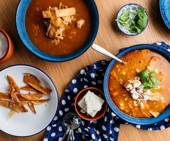 Bowls of tortilla soup with toppings; cilantro, tortilla strips, feta, on a table with a blue polka dot cloth.