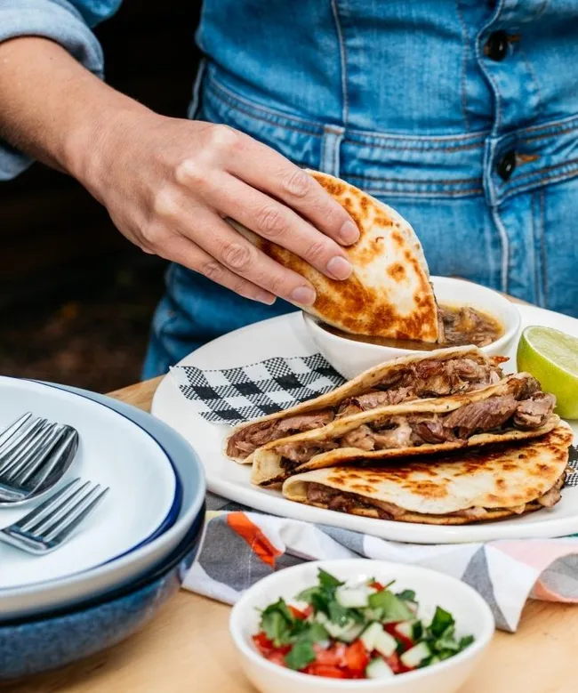 Person in denim jacket holding a birria taco over a bowl of broth, with plates and pico de gallo on the table.