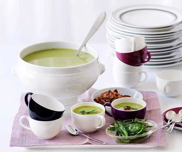 White soup tureen with green soup, cups, herbs, and spoons on a tray; stacked plates in the background.