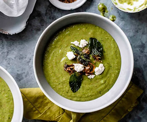 Creamy green soup in a white bowl, garnished with crumbled cheese and fresh mint leaves on a gray background.