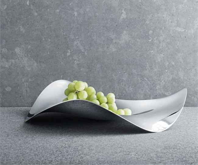 A modern, wavy metal tray holding green grapes on a gray textured surface.