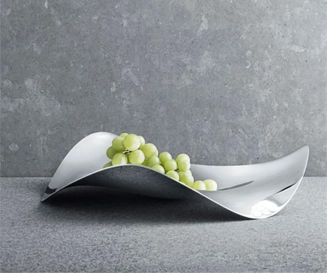 A modern, wavy metal tray holding green grapes on a gray textured surface.
