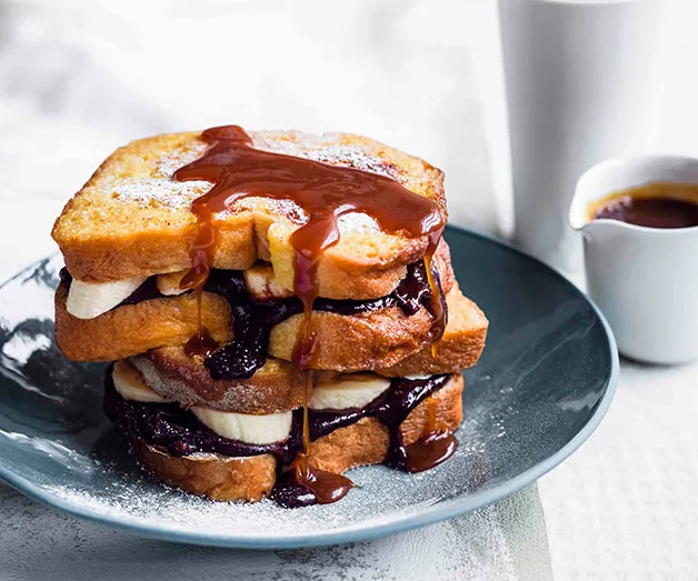 Stack of French toast with banana, chocolate spread, and caramel sauce on a blue plate, cup and jug in background.