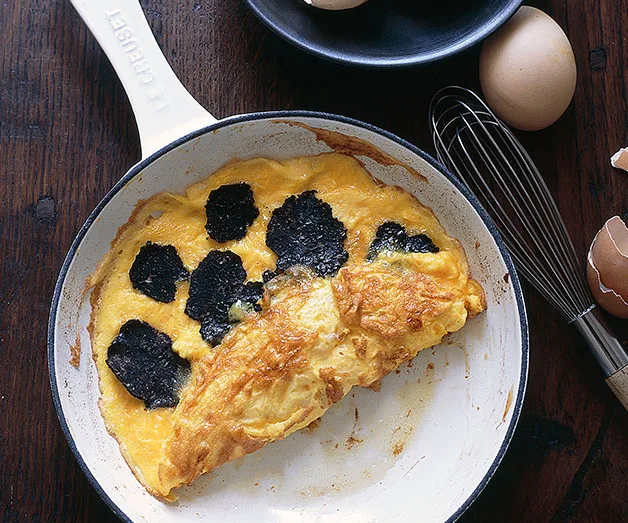 Truffle omelette in a white skillet on a wooden table with eggs and a whisk nearby.