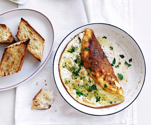 Omelette in a pan with herbs and melted ricotta, accompanied by slices of toasted bread on a plate.