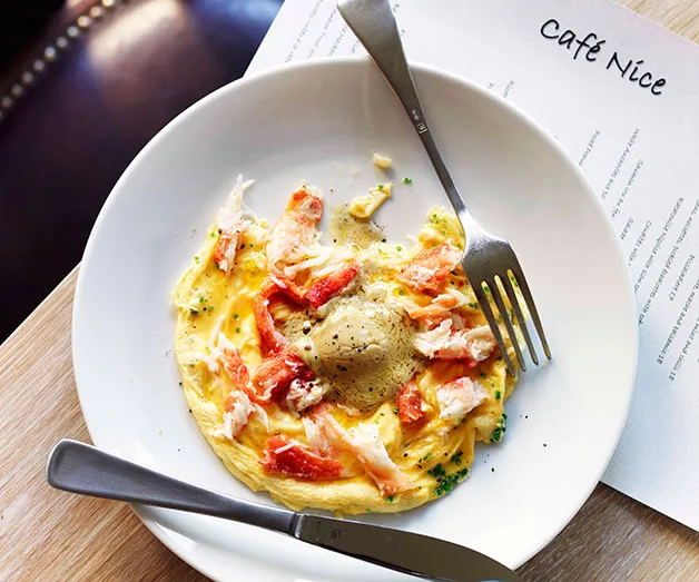 Plate of scrambled eggs with crab and truffle, fork and knife on the side, menu partially visible from Cafe Nice.