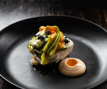 Plate of gourmet food with zucchini flowers, caviar, and sauce on a black dish.