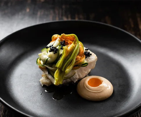 Plate of gourmet food with zucchini flowers, caviar, and sauce on a black dish.