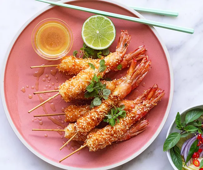 Crispy prawns on skewers with herbs, lime, and dipping sauce on a pink plate, accompanied by chopsticks.