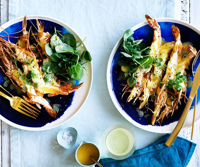 Grilled prawns with cheese and greens on blue plates, accompanied by gold cutlery on a light blue tablecloth.