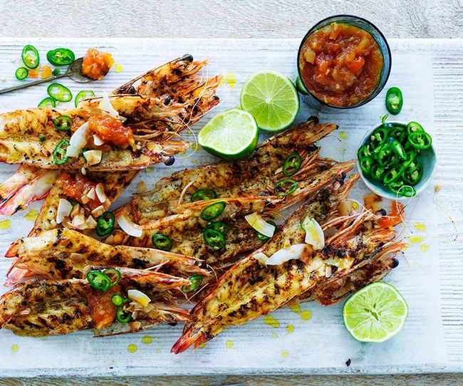 Grilled prawns topped with chili and salsa, served with lime halves and extra chili slices on a white platter.