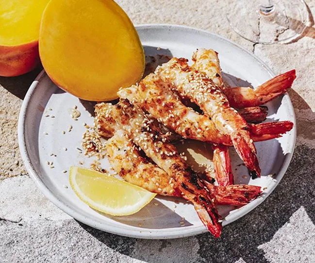 Grilled prawns with sesame seeds on a plate, served with lemon wedge and mango halves.