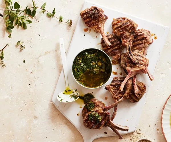 Grilled lamb chops on a white board with a bowl of green herb sauce and a spoon, garnished with fresh herbs.
