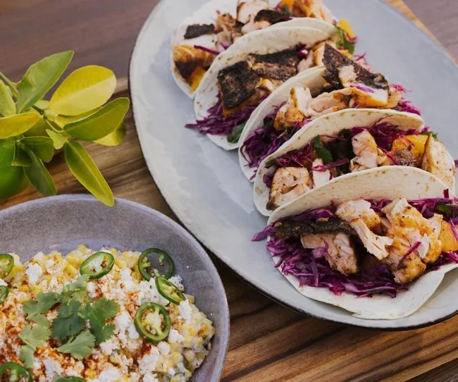Four fish tacos with cabbage and a bowl of elote topped with cheese, cilantro, and jalapeños, served on a wooden table.