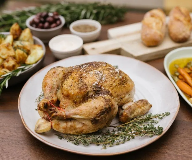 Roasted chicken on a plate with herbs, surrounded by sides of potatoes, bread, and carrots.