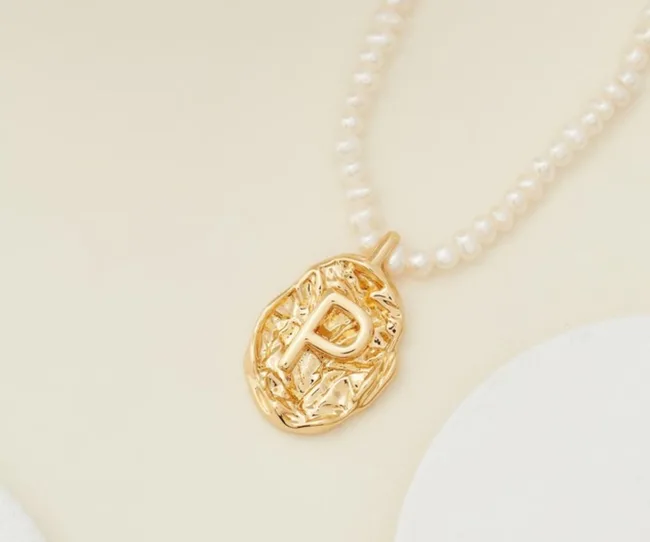Gold pendant with the letter "R" on a pearl necklace, set against a soft background.