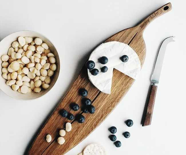 Cheese on a wooden board with blueberries, a cheese knife, and a bowl of macadamia nuts nearby.