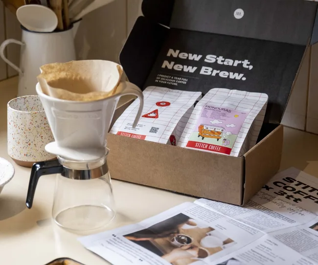 Coffee setup with a dripper and Stitch Coffee box labeled "Honduras," on a countertop with papers and a mug.