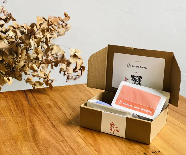 Open cardboard box on wooden table with coffee sample labeled "Ethiopia - Girma Sintayehu" and dried flowers nearby.