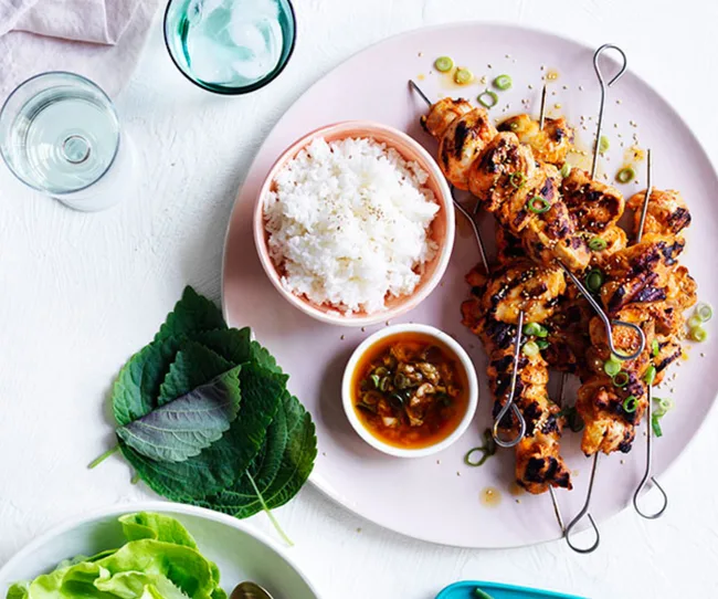 Grilled chicken skewers with rice and dipping sauce on a pink plate, accompanied by fresh leaves and a glass of water.