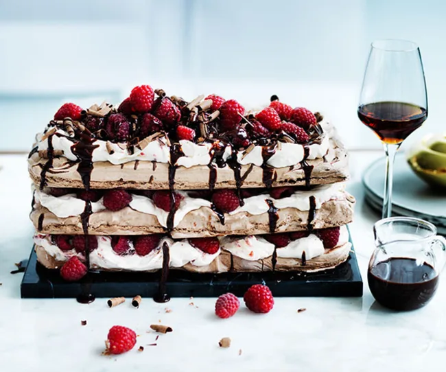 Layered cake with chocolate, cream, and raspberries, drizzled with sauce, on a marble table with a glass of red wine.