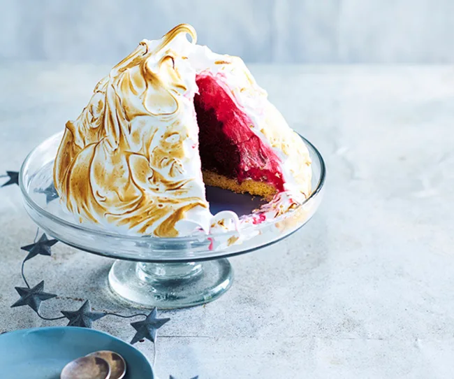 Baked Alaska dessert on glass stand, golden-browned meringue with a slice showing red filling and biscuit base.