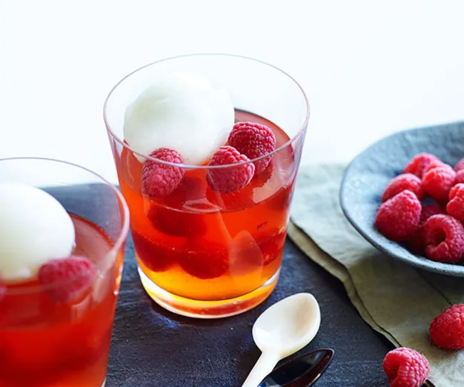 Raspberry dessert in a clear glass, topped with white sorbet, next to a bowl of fresh raspberries on a slate surface.