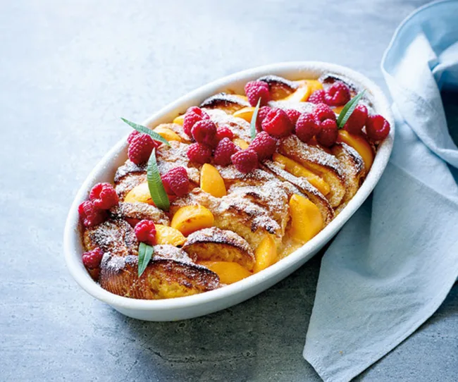 Fruit tart with raspberries and peaches in a white dish, garnished with powdered sugar and herbs, on a gray surface.