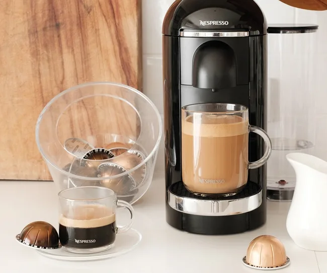 Nespresso machine with coffee cup, empty pods container, and a wooden cutting board on a kitchen counter.