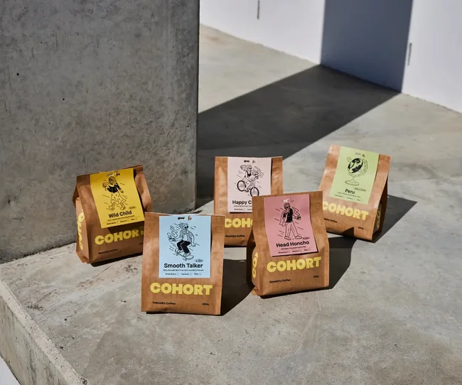 Five colorful Cohort coffee bags on a concrete surface, each with a unique design and label.
