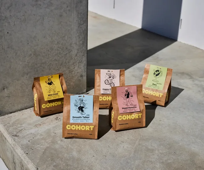 Five colorful Cohort coffee bags on a concrete surface, each with a unique design and label.