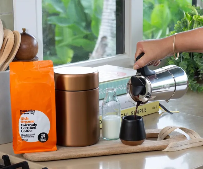 A person pours coffee from a silver pot into a black cup beside coffee bag, jar, milk bottle, and books on a countertop.