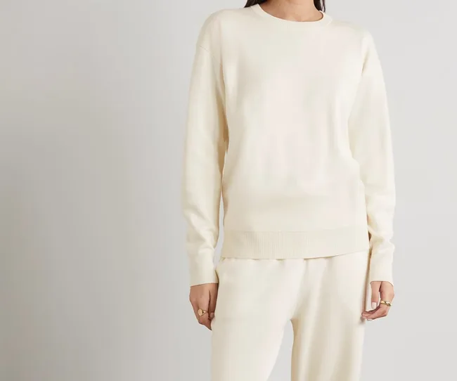 Woman in a cream sweater and pants, standing against a light background.