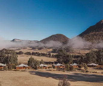 Rustic cabins in a valley with low-lying mist, surrounded by trees and mountains under a clear blue sky.