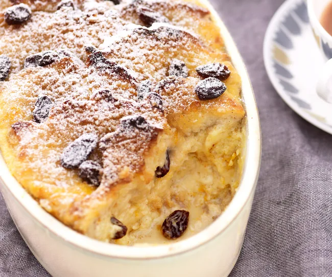 Bread pudding with raisins topped with powdered sugar in a white dish on a gray cloth.
