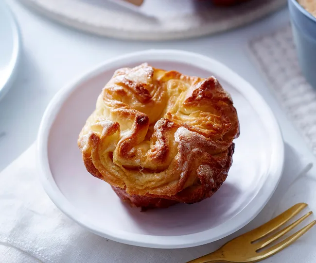 Golden-brown muffin on a white plate, with flaky, layered texture and crispy edges, beside a gold fork.