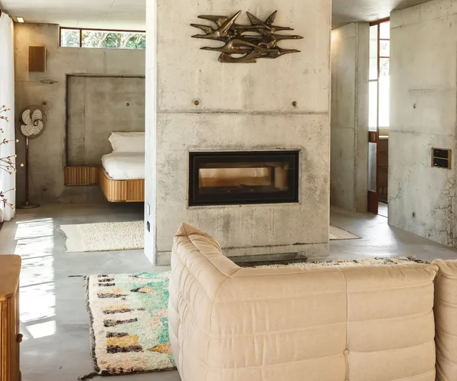 Modern living area with concrete walls, a beige couch, wall art, fireplace, and a bed in the adjacent space.